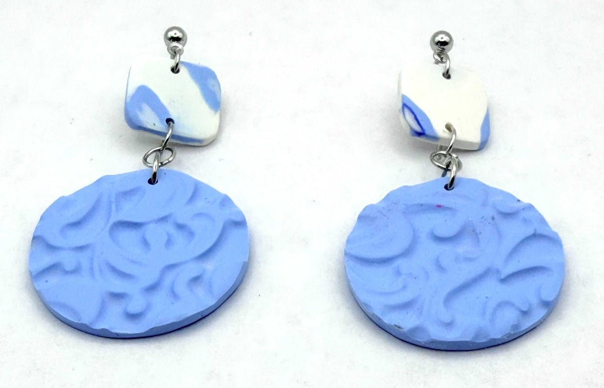 A pair of handmade drop earrings featuring textured light blue circular discs and white square charms with blue swirls, suspended from silver posts on a white background.