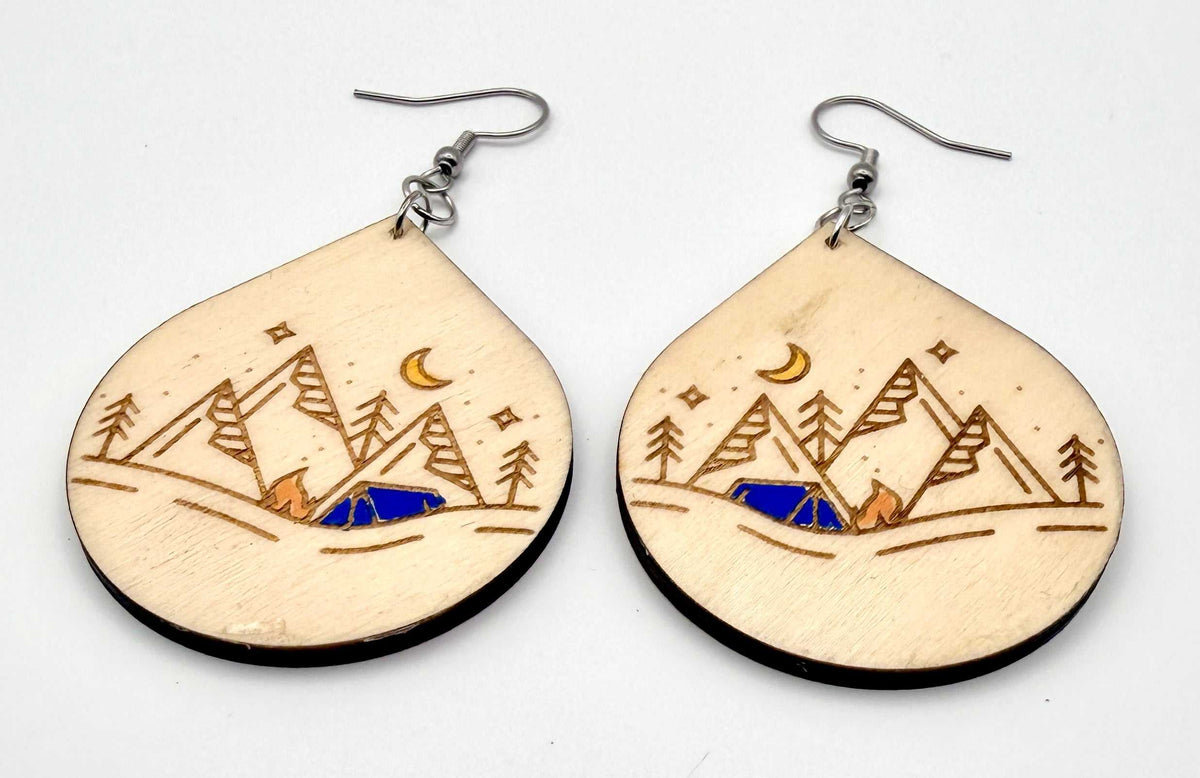 A pair of teardrop-shaped wooden dangle earrings with a laser-engraved mountain, moon, stars, and campfire design, shown on a plain white background.