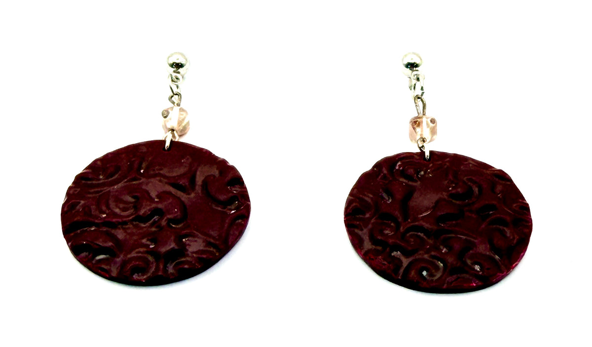 A pair of drop earrings featuring large, maroon circle discs with an embossed floral pattern, suspended from wire hooks with clear beads on a white background.