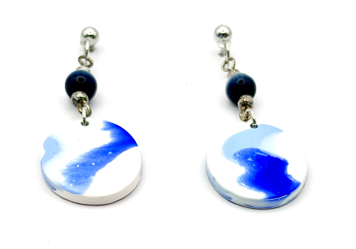 A pair of handmade dangle earrings featuring round white and blue marbled pendants, a dark blue bead, and silver-tone posts on a white background.