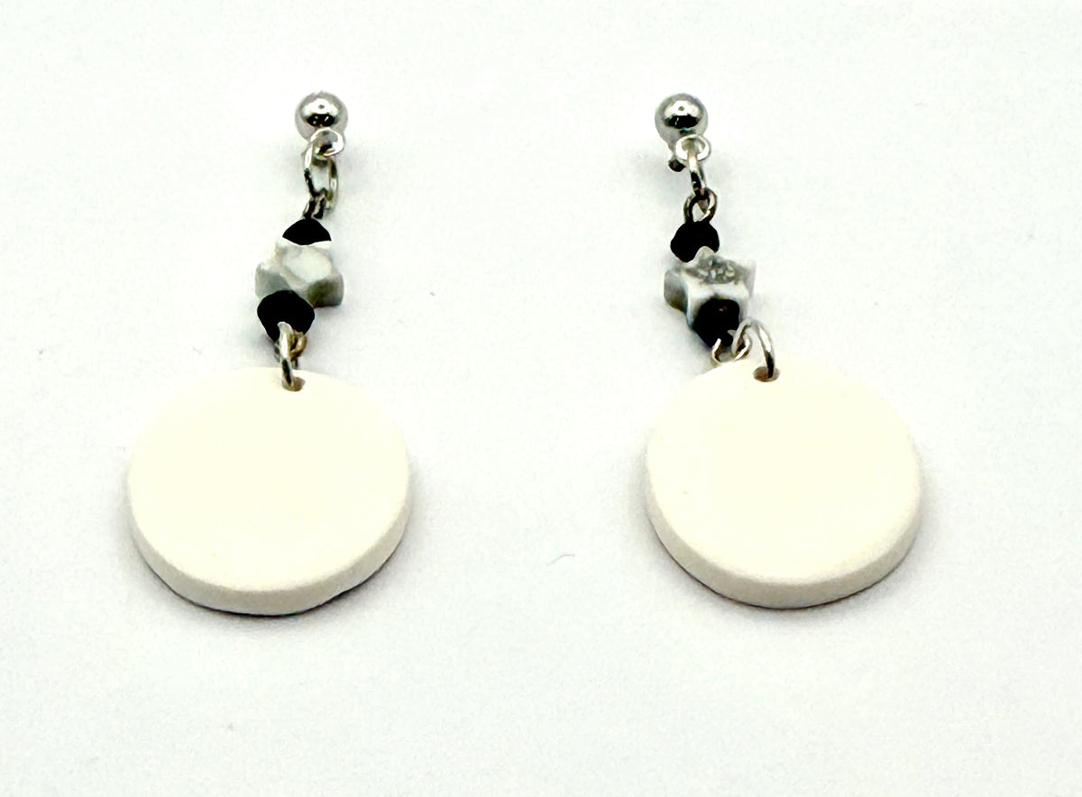 A pair of minimalist dangle earrings featuring large, flat circular disc charms in an off-white color, suspended from silver-toned posts with small black and marbled beads, set against a plain white background.