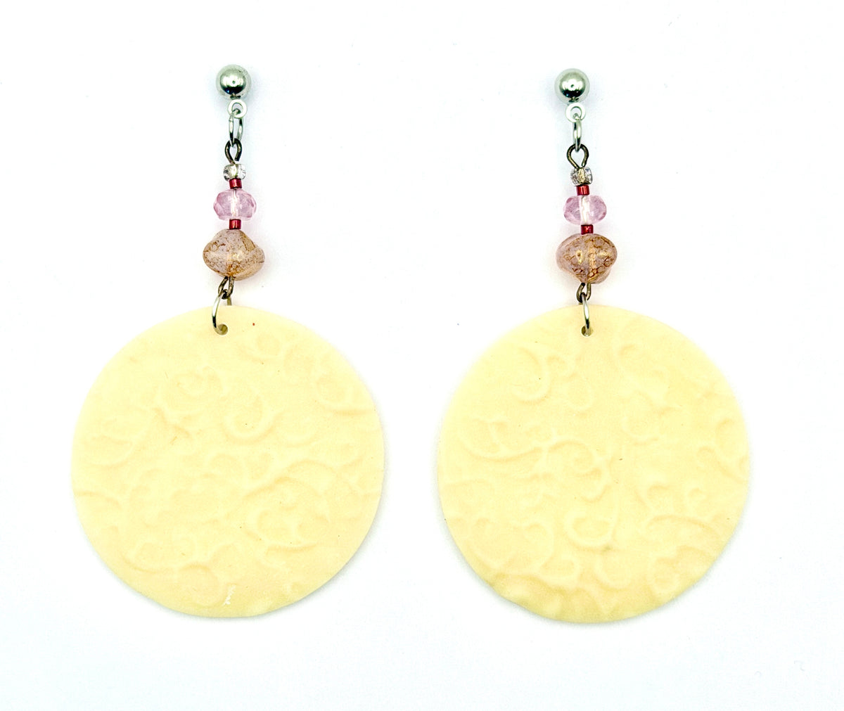 A pair of handmade dangle earrings featuring large, round, pale yellow disc charms with an embossed floral pattern, suspended from silver posts with pink faceted beads.