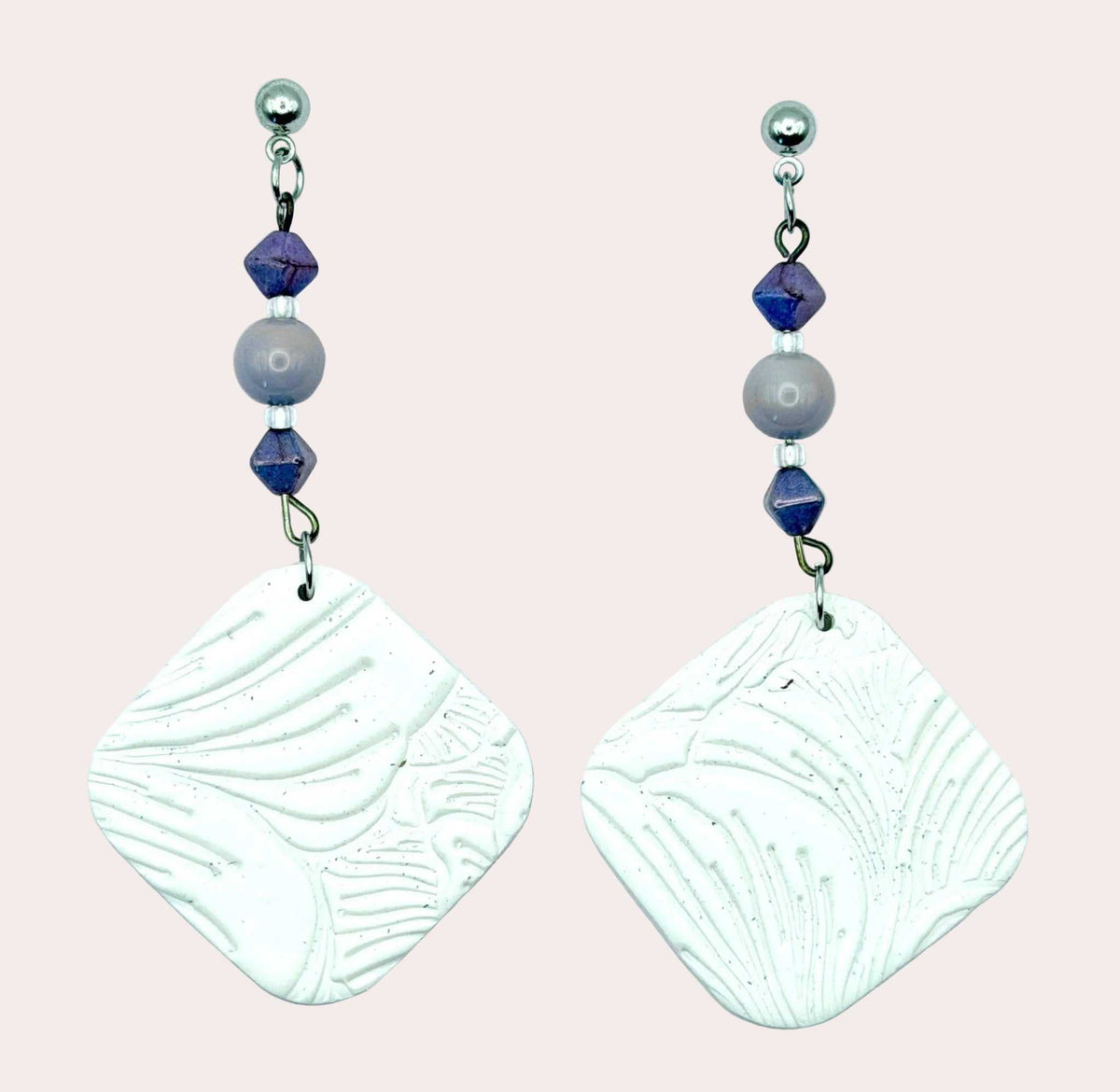 A pair of handmade dangle earrings featuring textured white square charms with leaf-like embossed patterns, suspended from silver hooks with purple and gray beads, set against a light blue background.