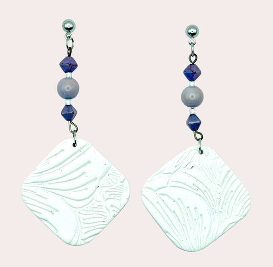 A pair of handmade dangle earrings featuring textured white square charms with leaf-like embossed patterns, suspended from silver hooks with purple and gray beads, set against a light blue background.