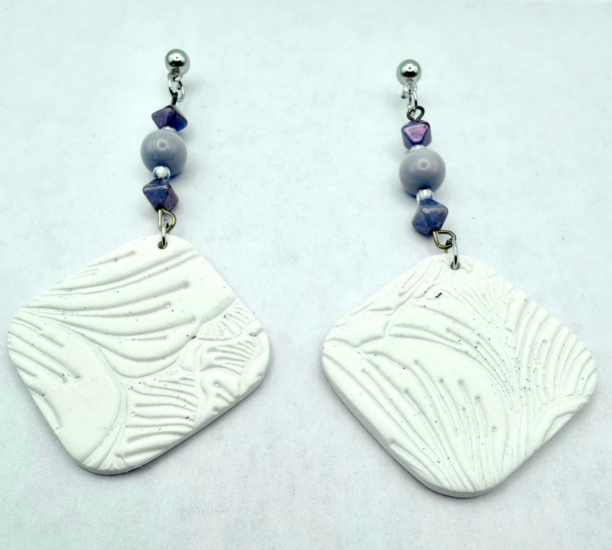 A pair of handmade dangle earrings featuring textured white square clay pendants with leaf-like embossed patterns, suspended from silver-toned posts with lavender and gray beads, set against a plain light gray background.
