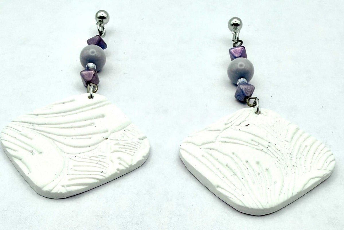 A pair of handmade dangle earrings featuring textured white clay discs with leaf-like embossed patterns, suspended from silver-toned posts with lavender and gray beads, set against a plain white background.