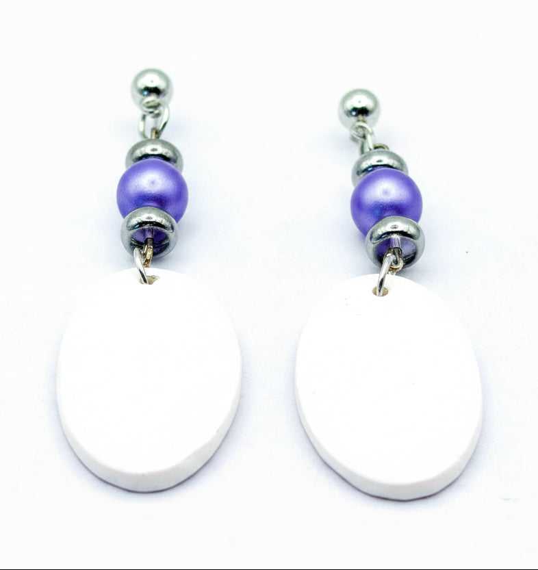 A pair of handmade dangle earrings featuring large, flat white oval charms, a small purple bead, and silver-toned metal spacers and posts, displayed on a light gray background.