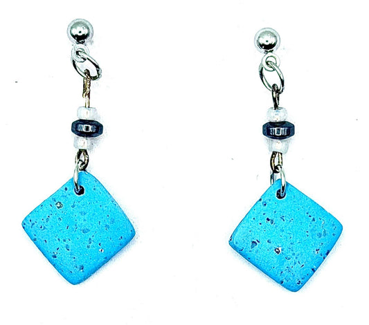 A pair of drop earrings featuring square-shaped, speckled turquoise blue ceramic pendants hanging from silver-tone posts with small dark and white beads.