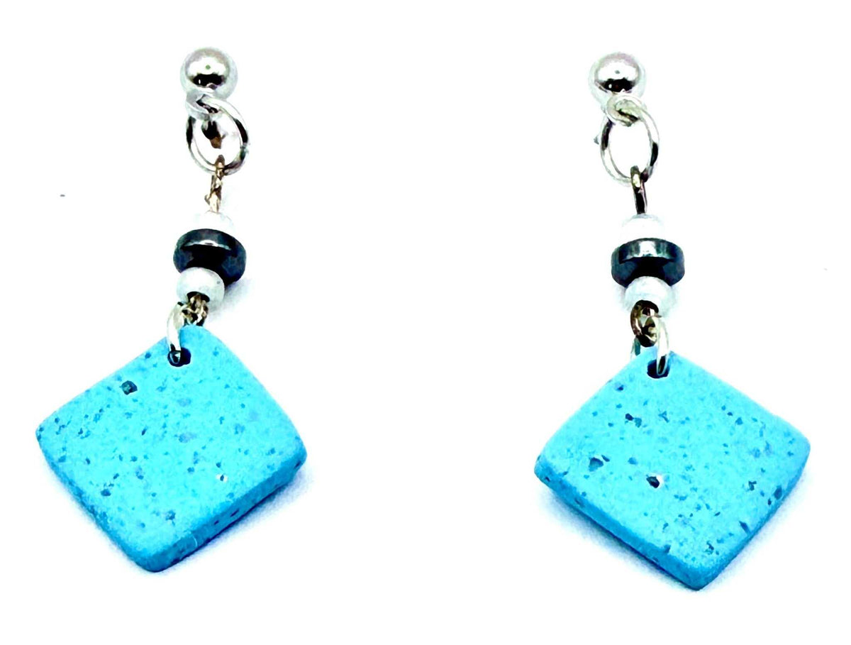 A pair of handmade dangle earrings featuring textured light blue square tiles, separated by a small black and white bead, hanging from silver-toned posts on a white background.