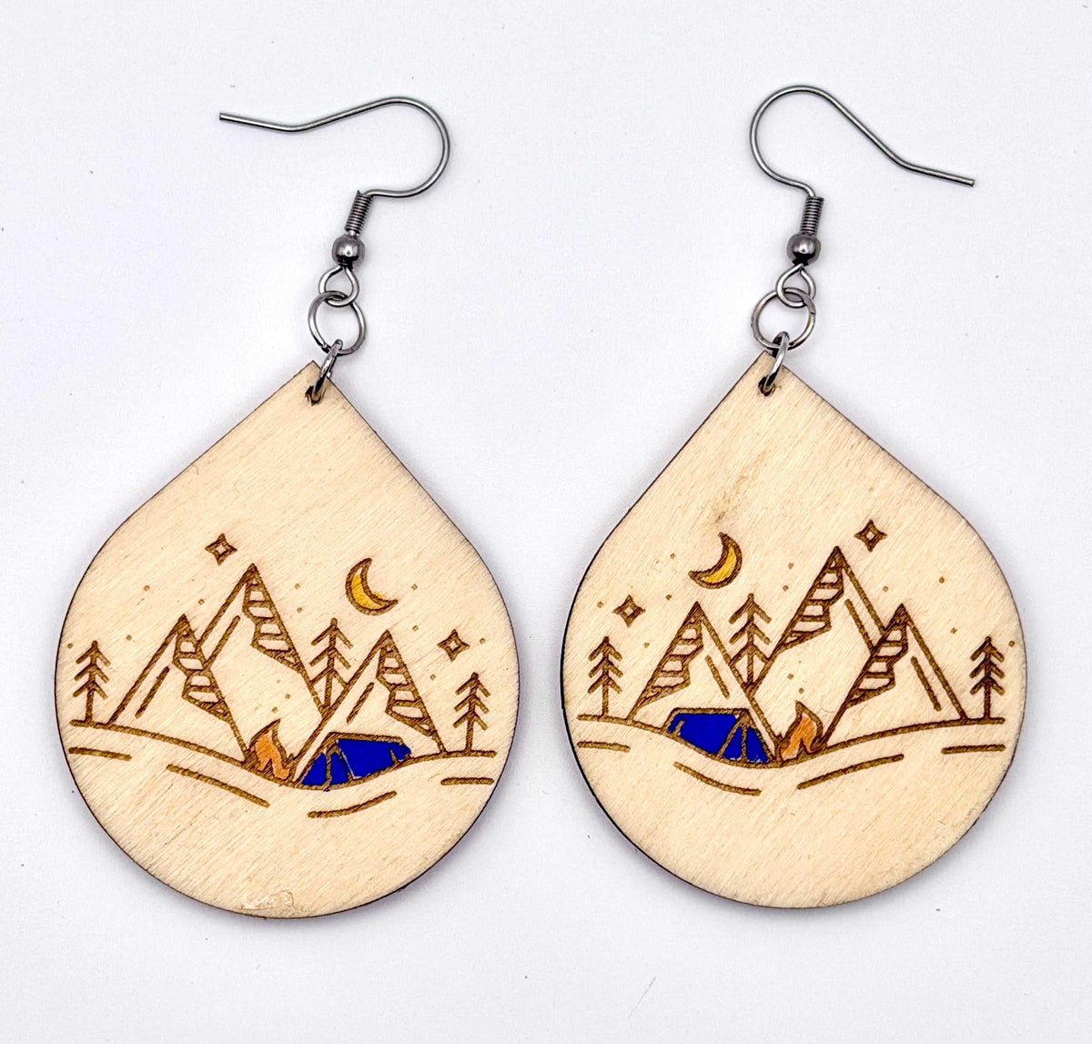 A pair of teardrop-shaped wooden drop earrings with laser-engraved mountain, trees, stars, and a crescent moon design, hanging from silver fishhook wires on a plain white background.