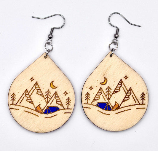 A pair of teardrop-shaped wooden drop earrings with laser-engraved mountain, trees, stars, and a crescent moon design, hanging from silver fishhook wires on a plain white background.