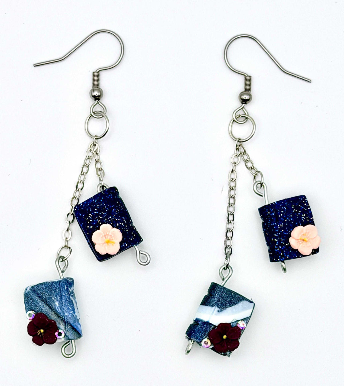 A pair of handmade dangle earrings on a white background. Each earring has a silver fishhook wire, a short silver chain, a dark blue glitter rectangular charm with a small pale pink flower, and a dangling blue-and-white marbled charm with a dark red flower.