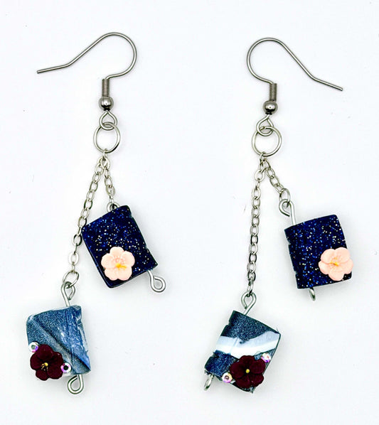 A pair of handmade dangle earrings on a white background. Each earring has a silver fishhook wire, a short silver chain, a dark blue glitter rectangular charm with a small pale pink flower, and a dangling blue-and-white marbled charm with a dark red flower.