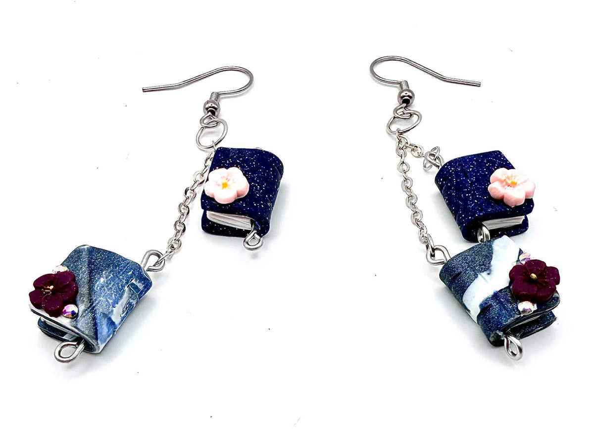 A pair of handmade dangle earrings featuring miniature book charms. The earrings include silver fishhook wires, a short silver chain, a dark blue glitter book charm with a small pink flower, and a blue-and-white marbled book charm with a dark red flower.