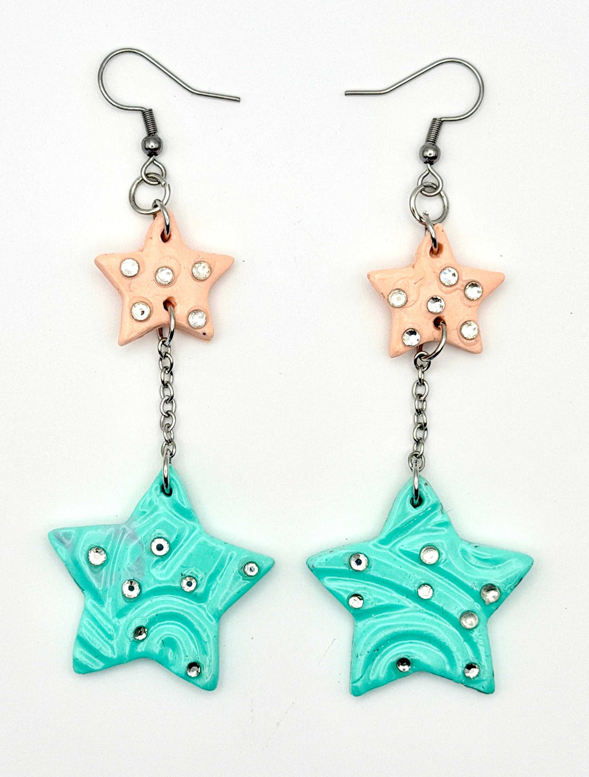 A pair of handmade dangle earrings featuring a peach star charm with rhinestones, connected by a silver chain to a larger turquoise star charm with swirl patterns and rhinestones, on a white background.
