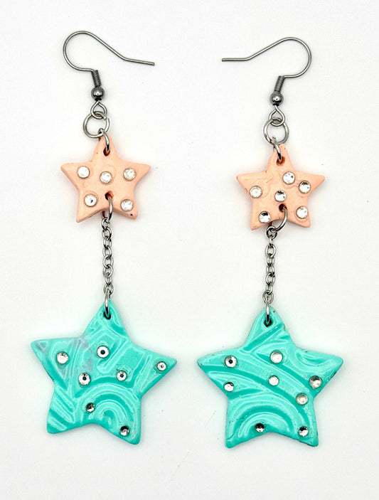 A pair of handmade dangle earrings featuring a peach star charm with rhinestones, connected by a silver chain to a larger turquoise star charm with swirl patterns and rhinestones, on a white background.