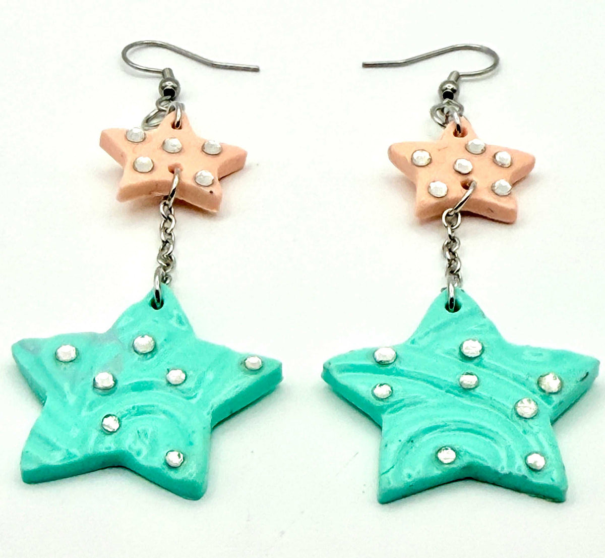 A pair of handmade dangle earrings featuring a peach star charm with silver rhinestones, hanging from a silver fishhook ear wire, with a turquoise star pendant below connected by a short silver chain.