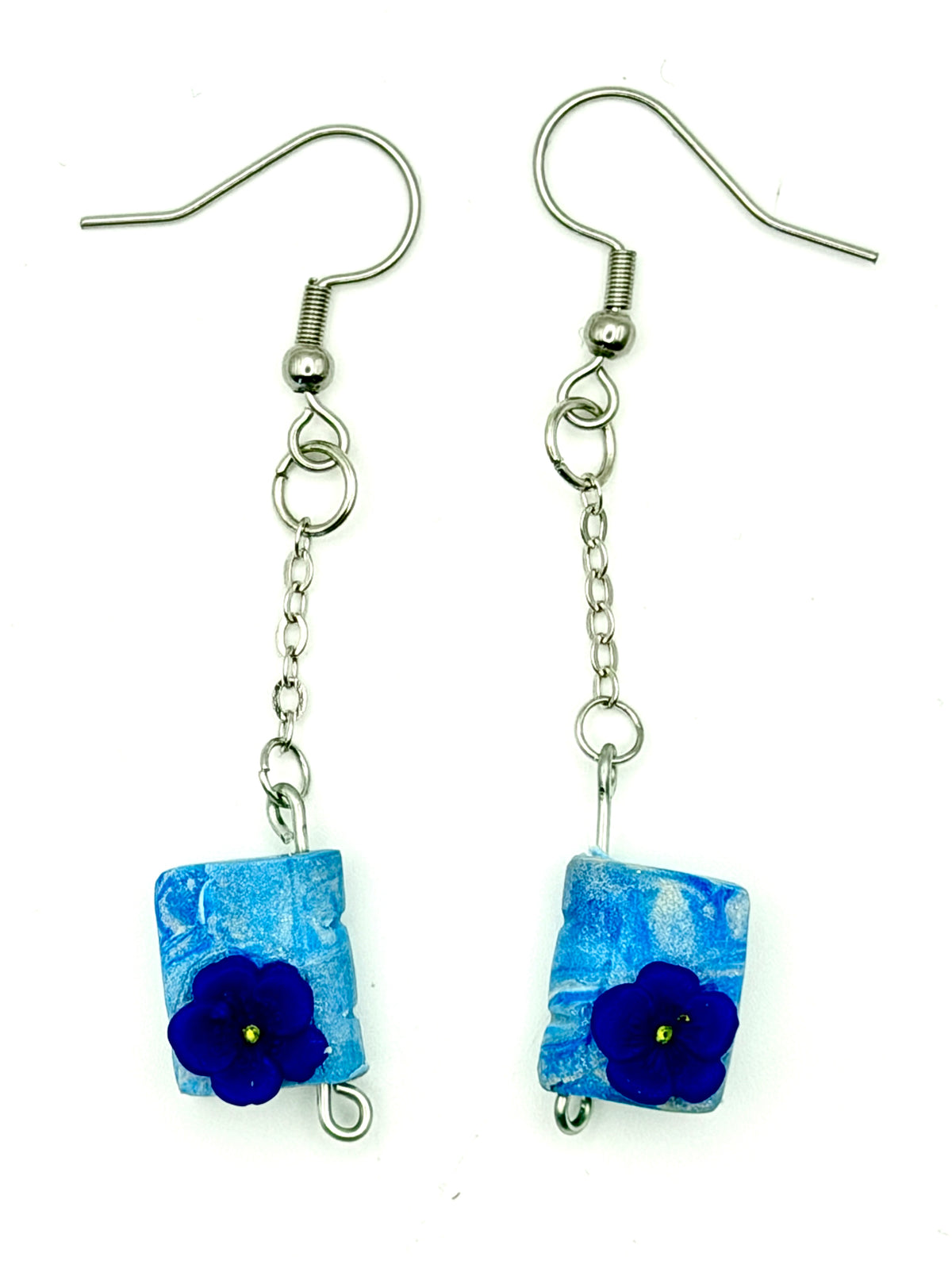 A pair of handmade dangle earrings featuring textured blue book-shaped pendants with a dark blue flower accent, suspended from silver-tone fishhook wires on a white background.