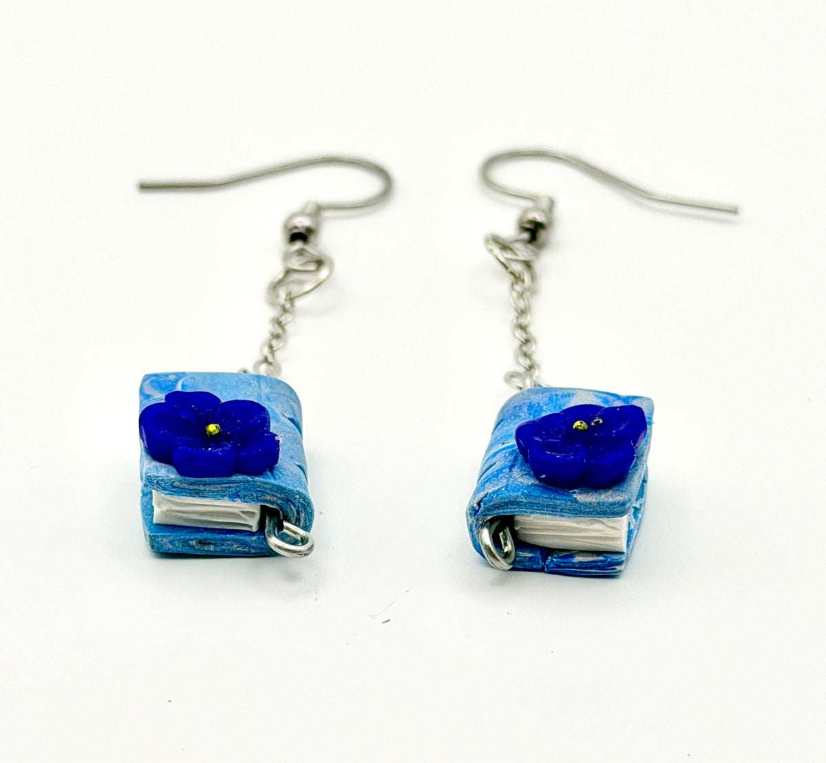 A pair of handmade dangle earrings featuring miniature blue books with a glossy blue flower charm on each, suspended from silver fishhook wires on a white background.