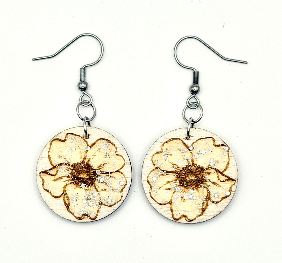 A pair of handmade dangle earrings featuring circular wooden discs engraved with a floral design and decorated with silver glitter, hanging from silver-tone fishhook wires on a plain white background.