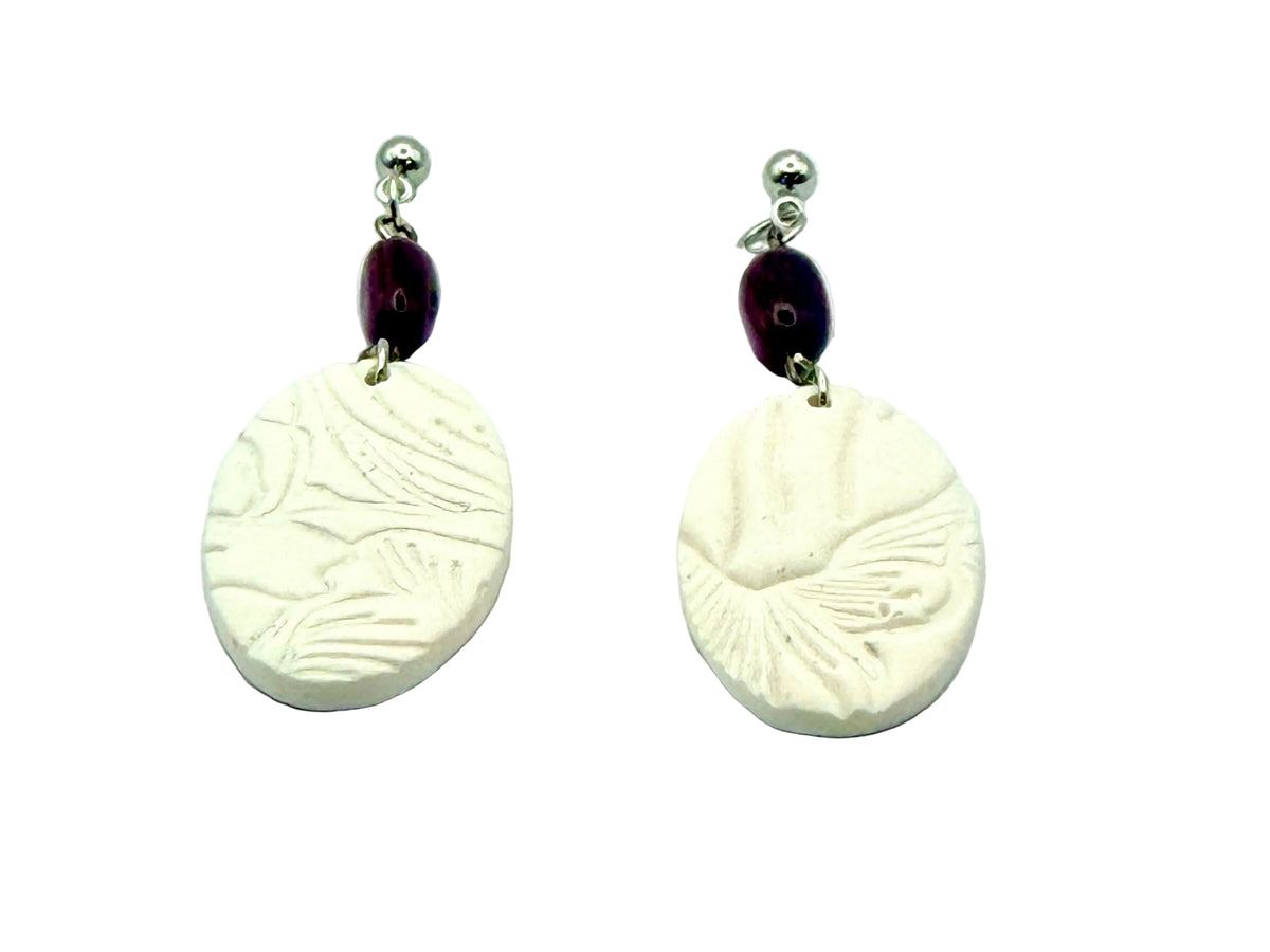 A pair of handmade dangle earrings featuring oval white clay pendants with embossed leaf patterns, connected by a dark purple bead and a silver-tone ball stud.