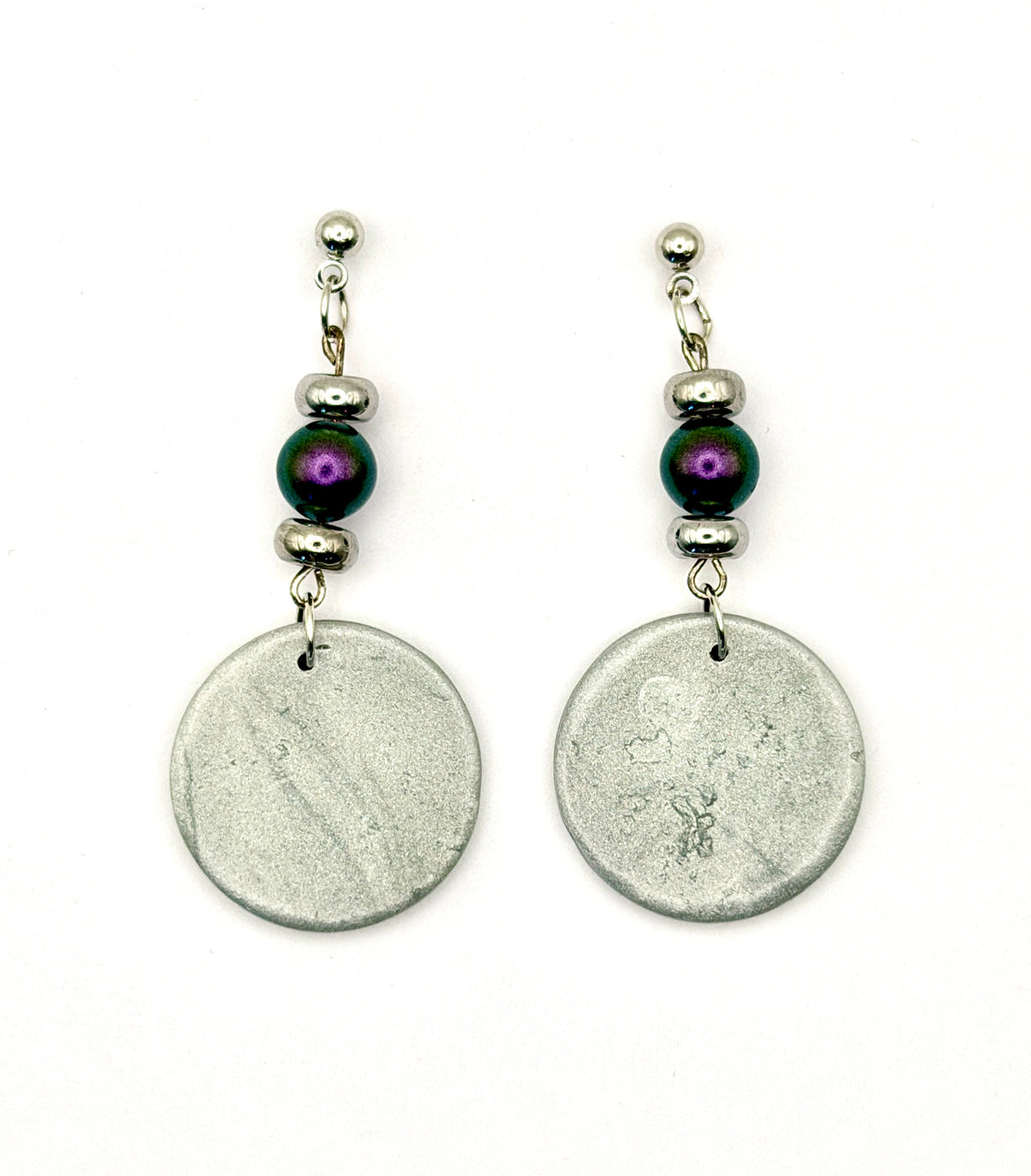 A pair of handmade dangle earrings featuring textured silver disc pendants, silver spacer beads, and a single dark purple iridescent bead, shown on a plain white background.