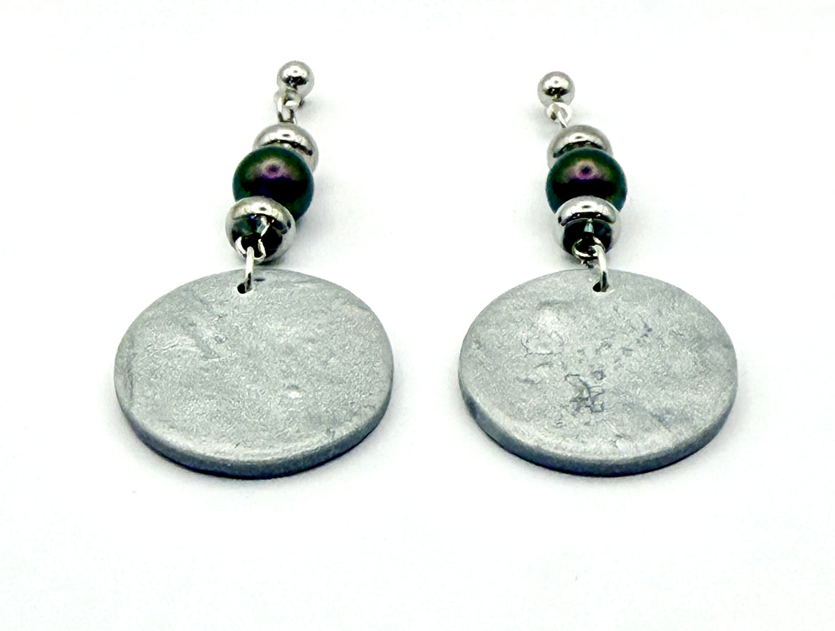 A pair of handmade dangle earrings featuring textured silver oval discs, dark purple beads, and silver spacer beads on a white background.