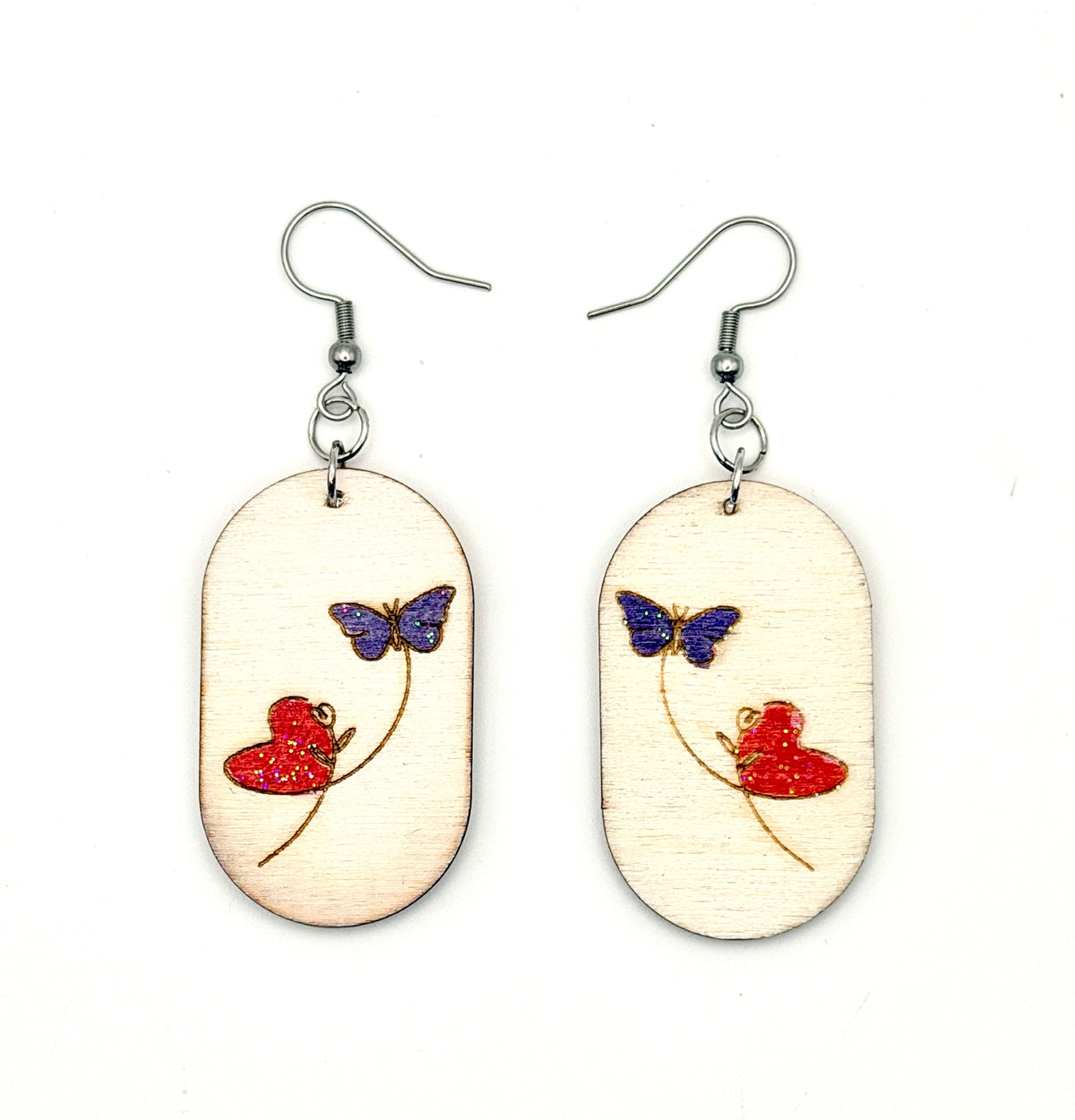 A pair of wooden dangle earrings with silver fishhook wires. The oval wooden charms are light beige wood with a hand-painted design featuring a red heart, and a purple butterfly.