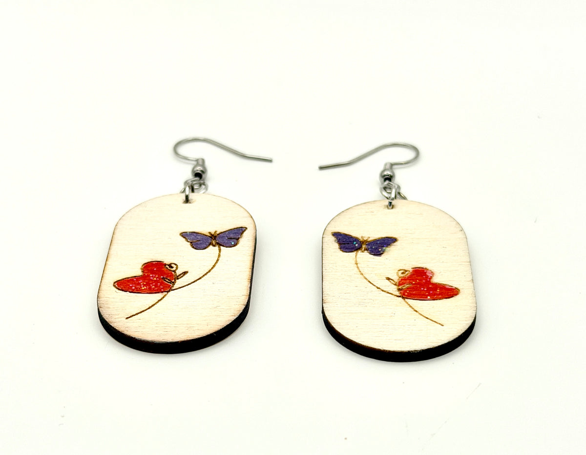 A pair of wooden dangle earrings with silver fishhook wires. The oval-shaped light wood earrings feature a hand-painted red heart and a purple butterfly design.