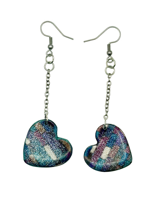 A pair of dangling heart-shaped earrings with a glittery finish in shades of blue, purple, and pink, suspended from silver-toned fishhook wires on a plain white background.