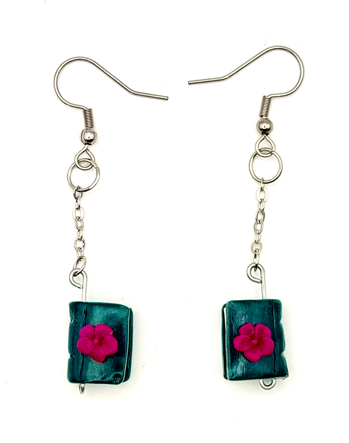 A pair of handmade dangle earrings featuring textured, dark teal-green polymer clay books with a small, vibrant magenta-pink flower attached to each, suspended from silver-tone fishhook wires on a plain white background.