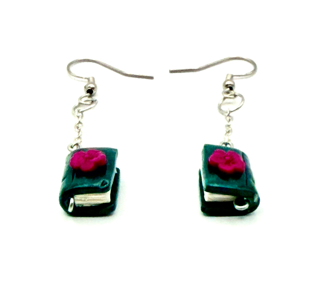 A pair of dangling earrings shaped like miniature dark green books with bright pink flower embellishments on the covers, suspended from silver fishhook wires against a plain white background.