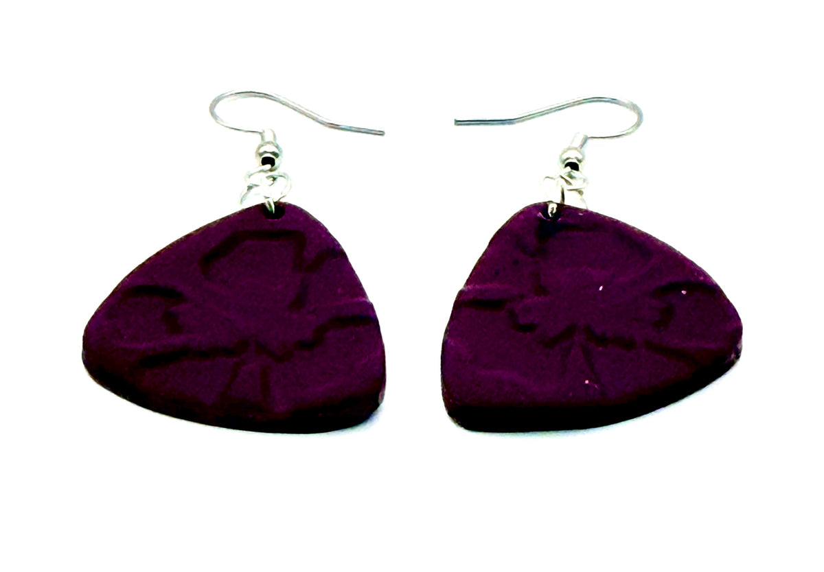 A pair of handmade dangle earrings featuring dark purple, guitar-pick-shaped beads with an embossed flower design, suspended from silver fishhook wires on a white background.