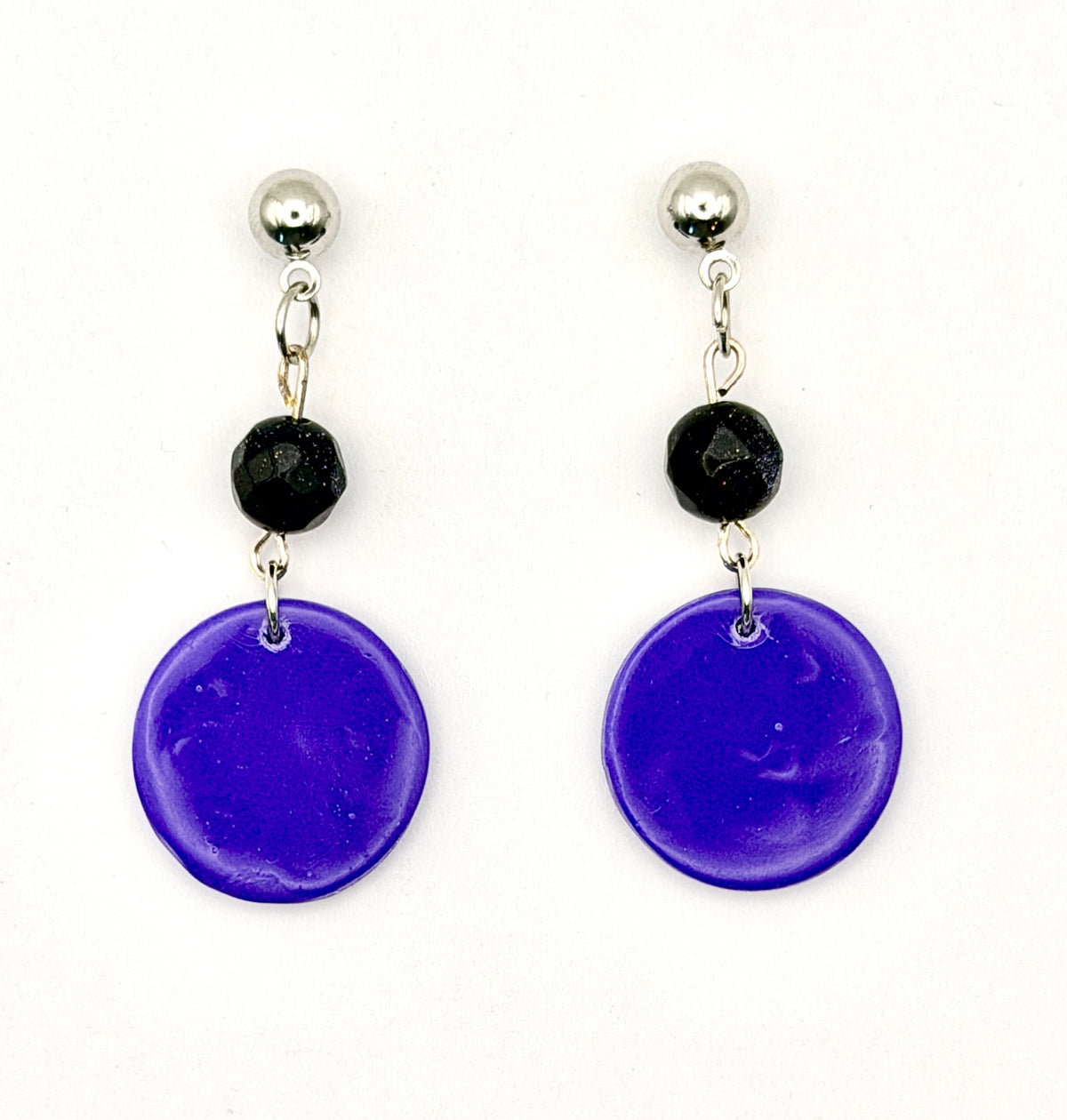 A pair of handmade dangle earrings featuring large, textured deep purple circular discs, a small black faceted bead, and silver-tone metal studs on a plain white background.