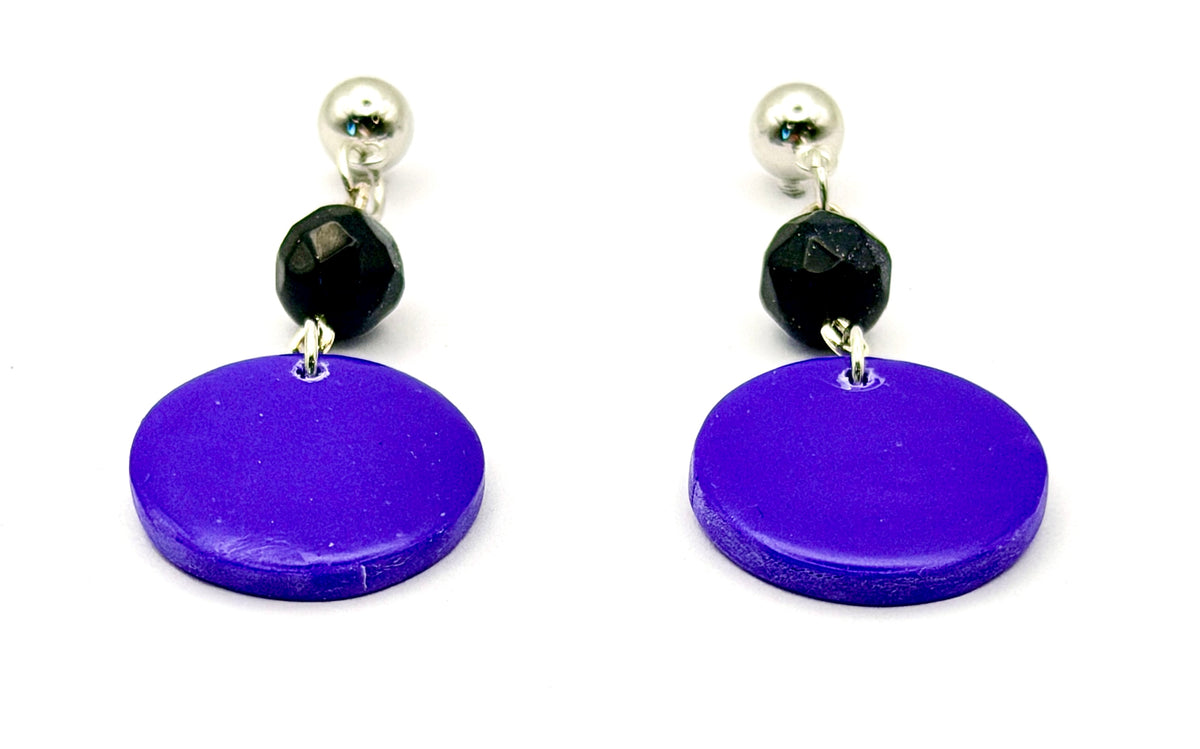A pair of drop earrings featuring large, round, vibrant purple disc charms, a small faceted black bead, and a polished silver ball stud at the top, shown against a plain white background.