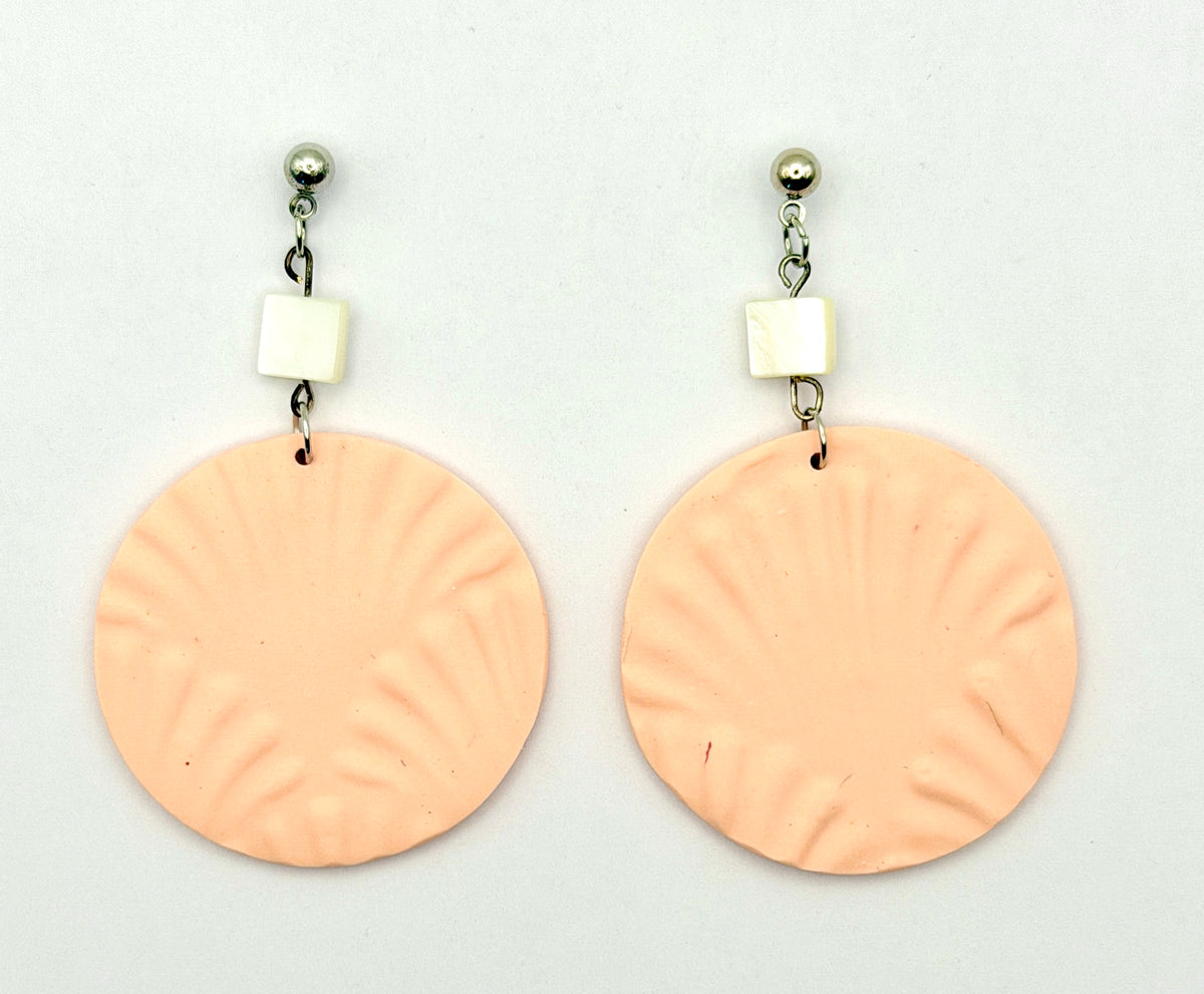 A pair of handmade dangle earrings featuring large, textured peach-pink circular discs with radial grooves, suspended from silver posts with small white square beads, shown on a plain white background.
