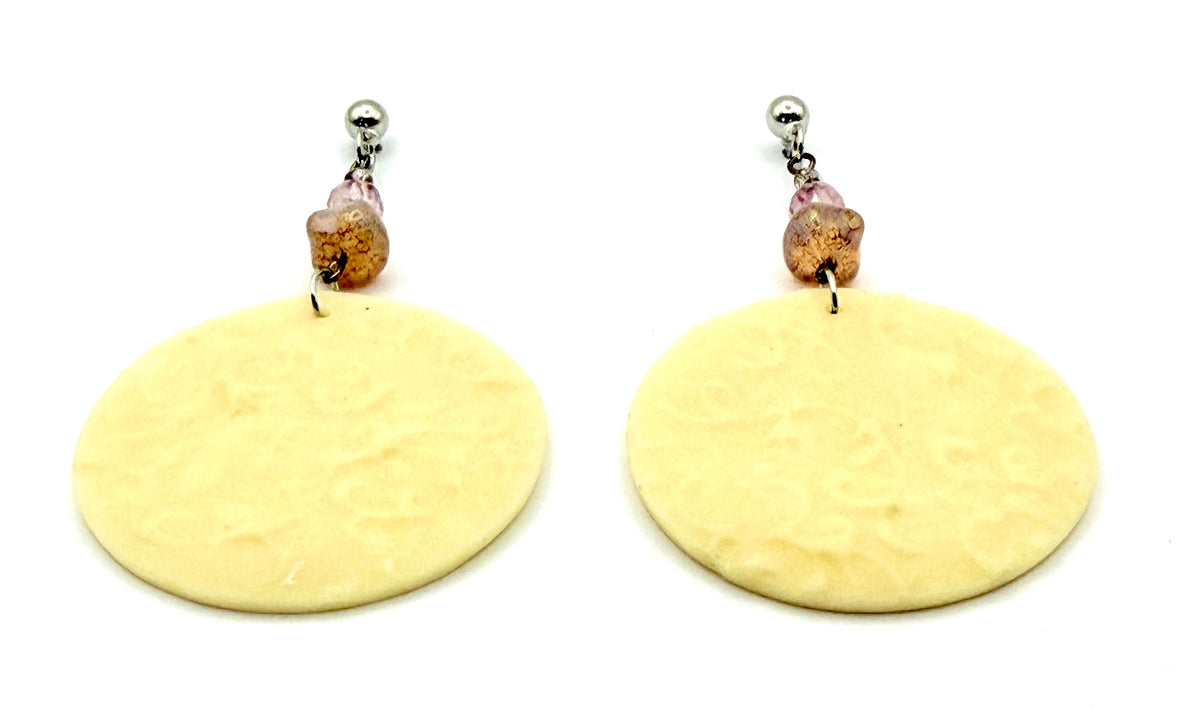 A pair of handmade dangle earrings featuring textured, oval-shaped cream-colored discs suspended from small pinkish-brown beads and silver posts, shown against a plain white background.